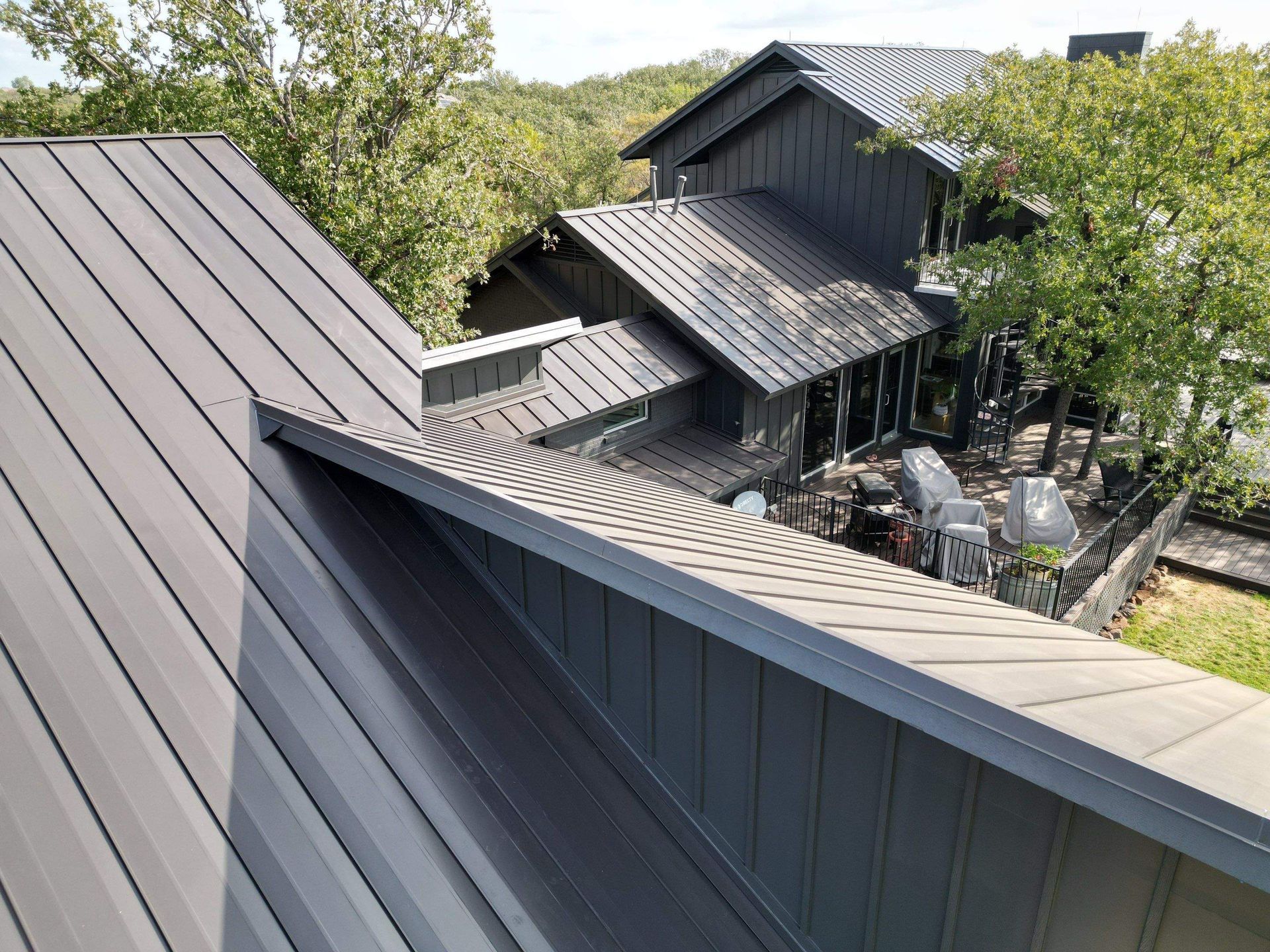 Rooftop view of a gray metal-clad house surrounded by green trees, seen from above.