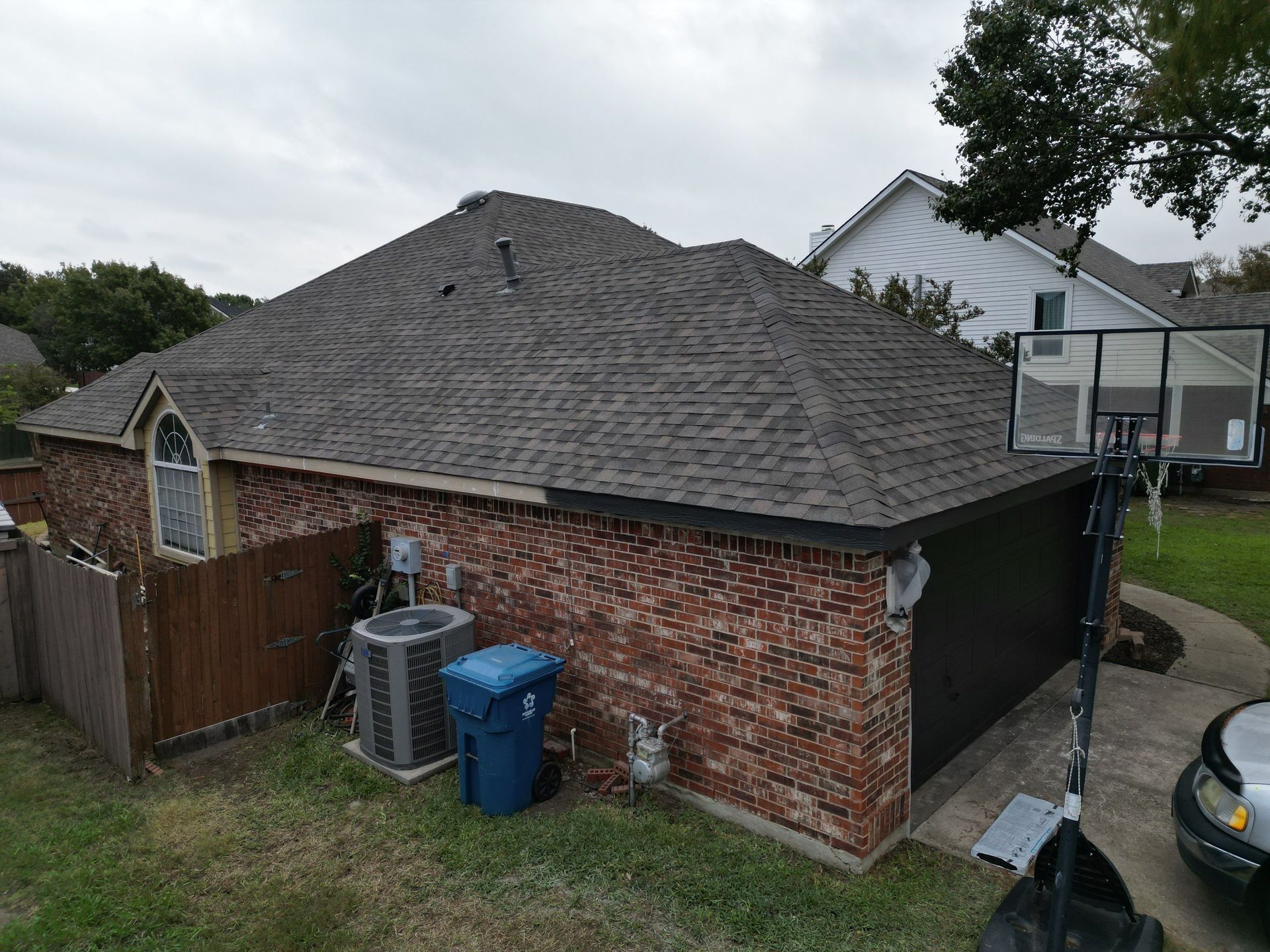 Aerial view of a brick house with a gray shingle roof, fenced yard, trash bins, and a parked car.