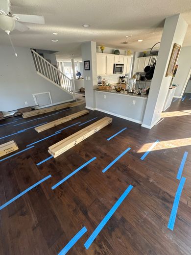 Living room under renovation with blue tape lines on dark wood floor and scattered lumber near staircase and kitchen