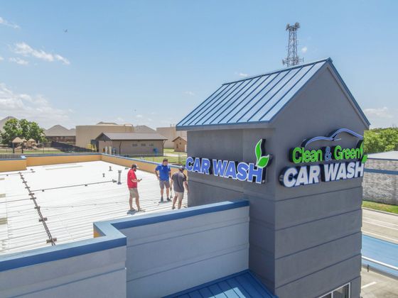 Outdoor car wash with a blue-roofed building and a few people standing on a sunlit roof deck.