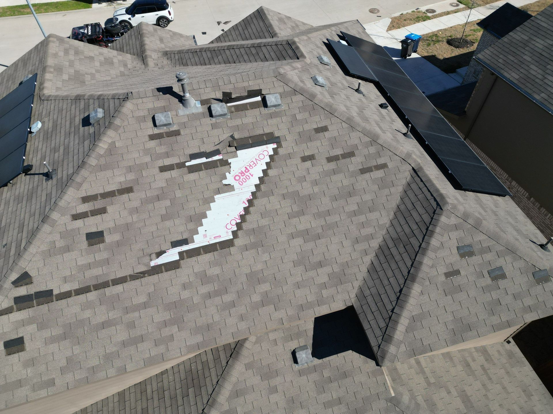 Aerial view of a gray shingled roof with a damaged patch and visible roof vents.
