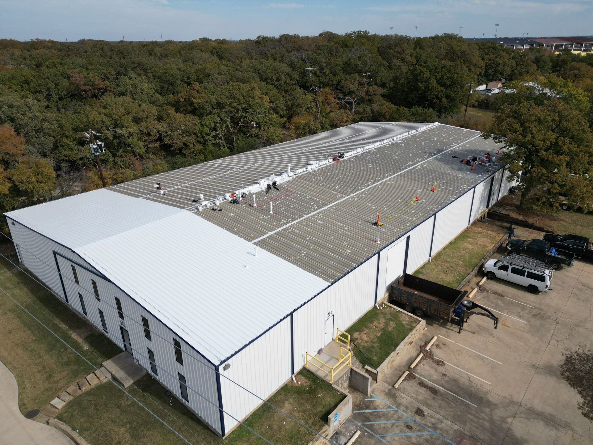 Aerial view of a large white warehouse with a curved roof, surrounded by trees and parked cars.