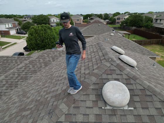 Person walking on a gray shingled rooftop with white roof vents in a suburban neighborhood.