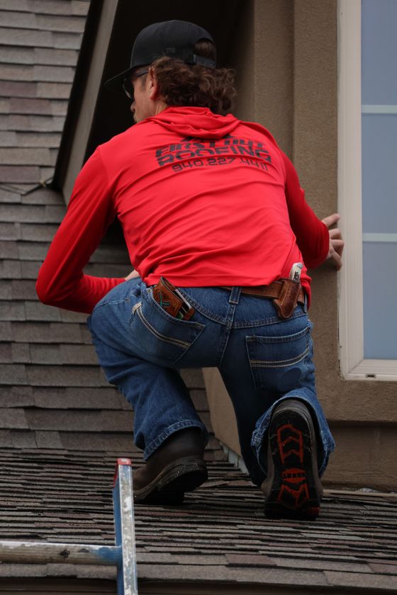 Worker in red shirt kneeling on a shingled roof near a window, using tools at the edge.