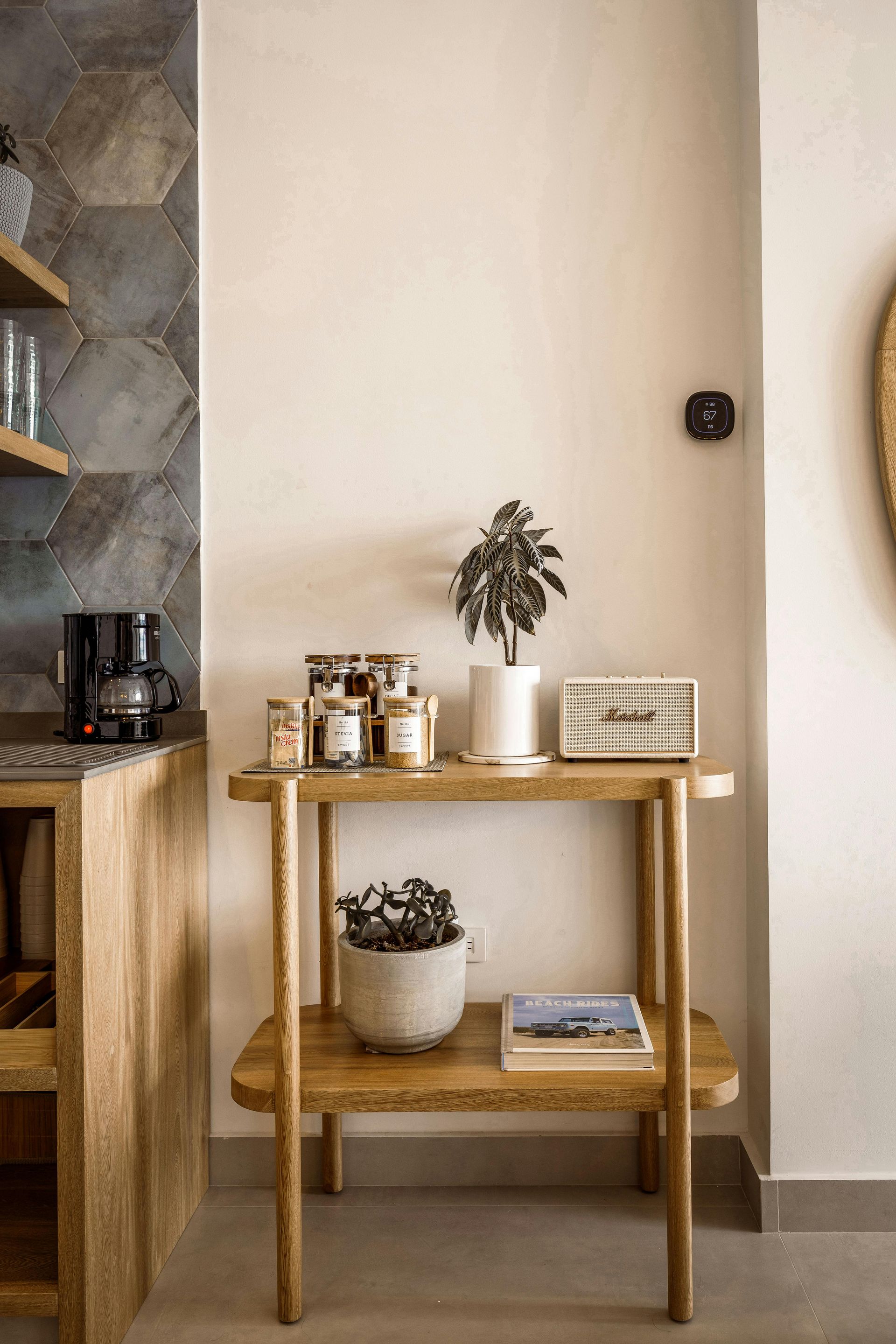 A two-tiered wooden side table holds plants, jars, a speaker, and a book against a white wall next to a tiled kitchen.