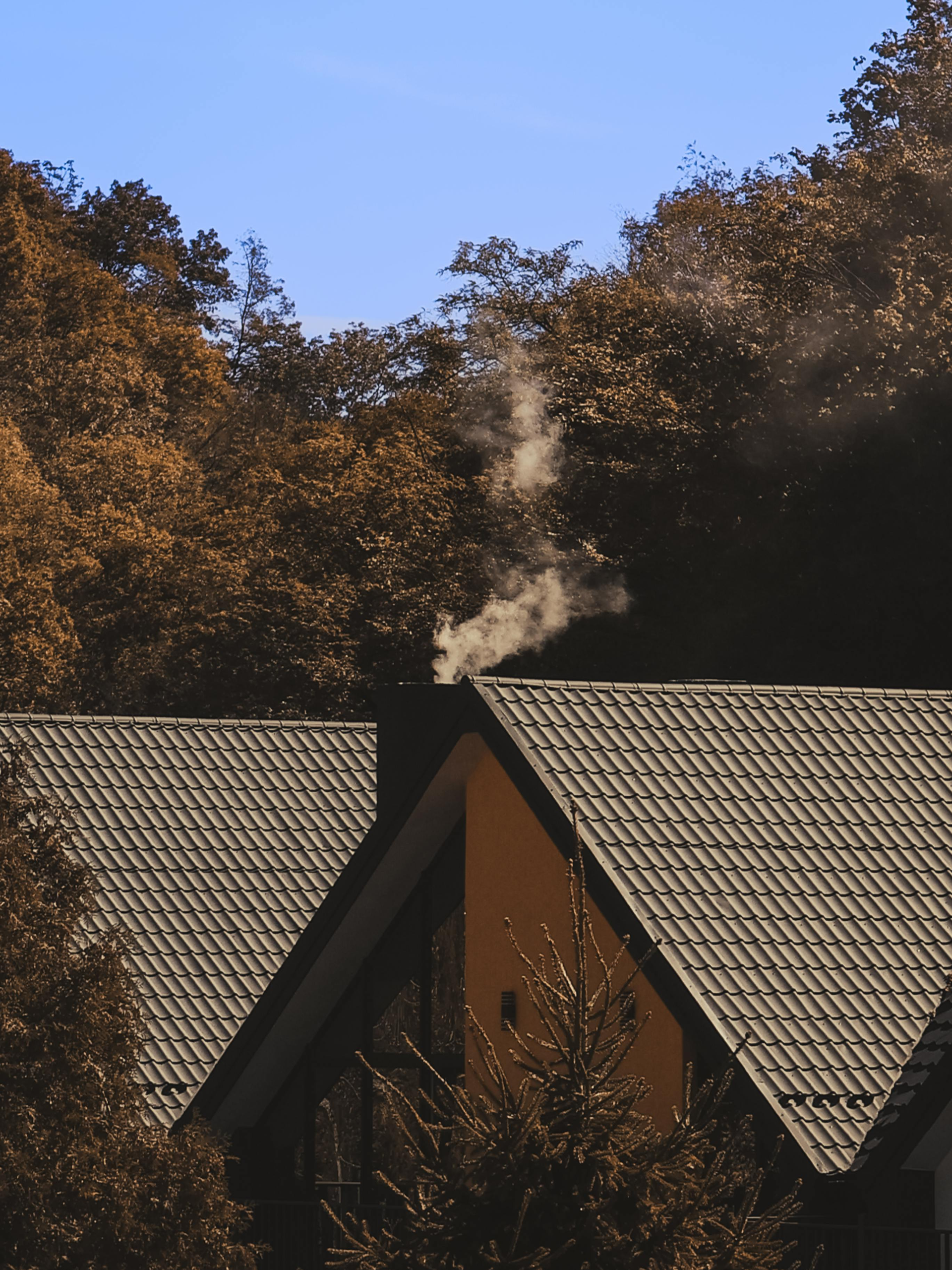 Smoke rising from the chimney of a house with a grey tiled roof, framed by autumn trees under a clear blue sky.