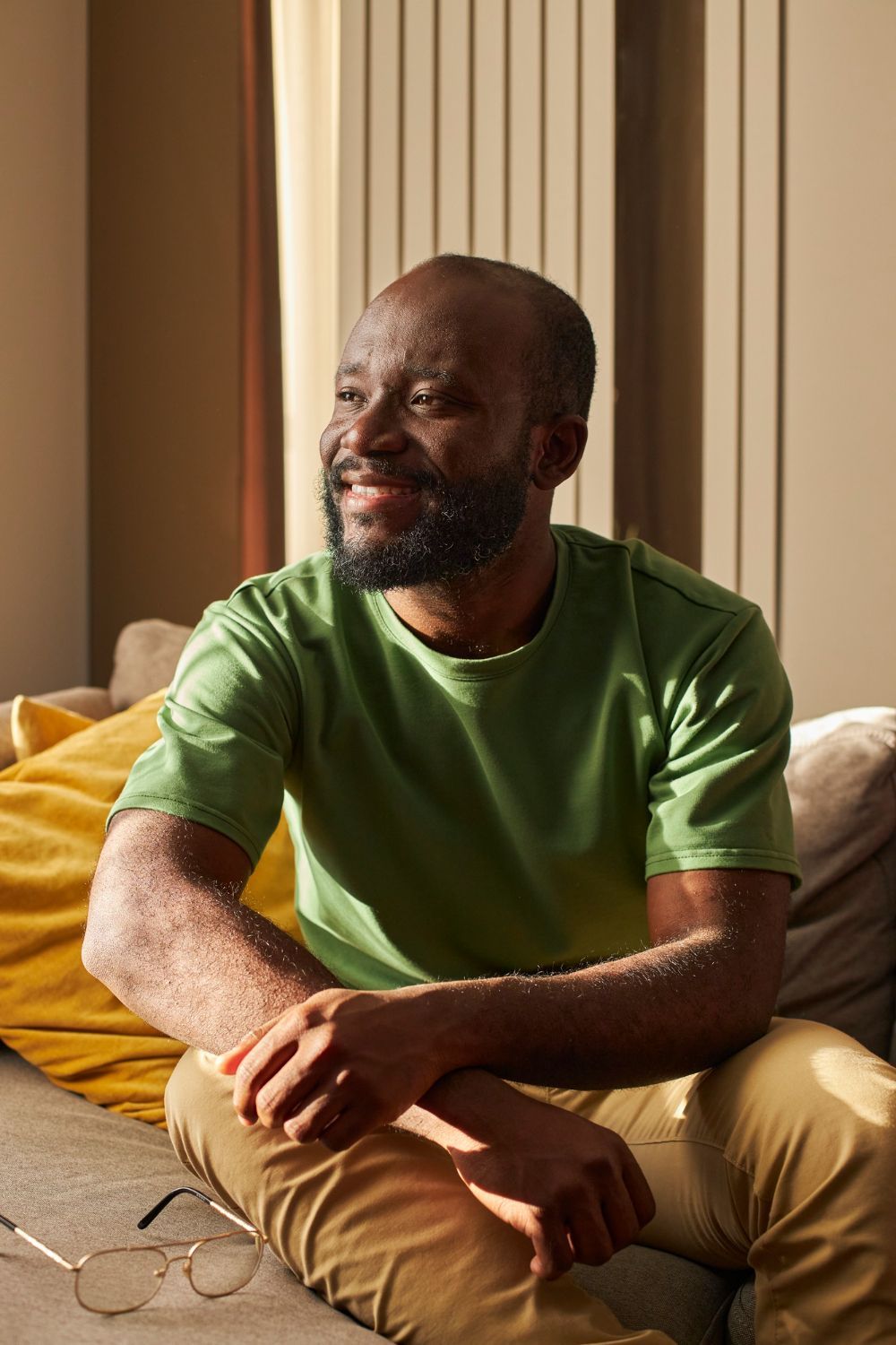 Man wearing green shirt and khaki pants sitting on a couch, smiling, sunlight.