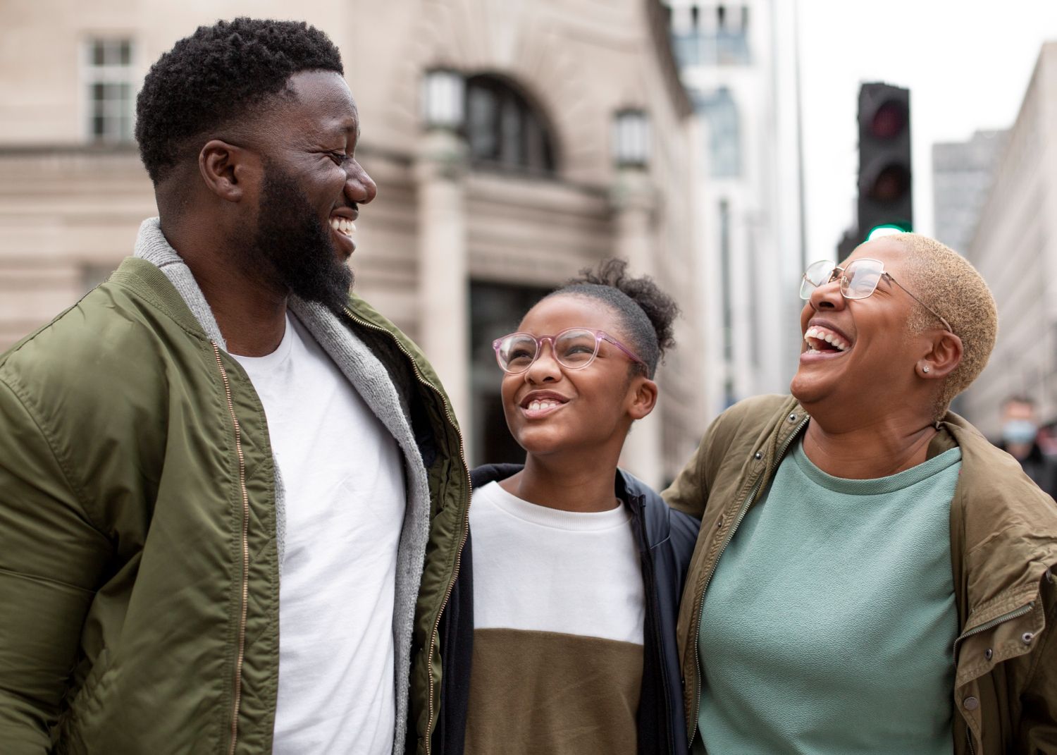 Family of three laughing together outdoors in front of a building with a traffic light.
