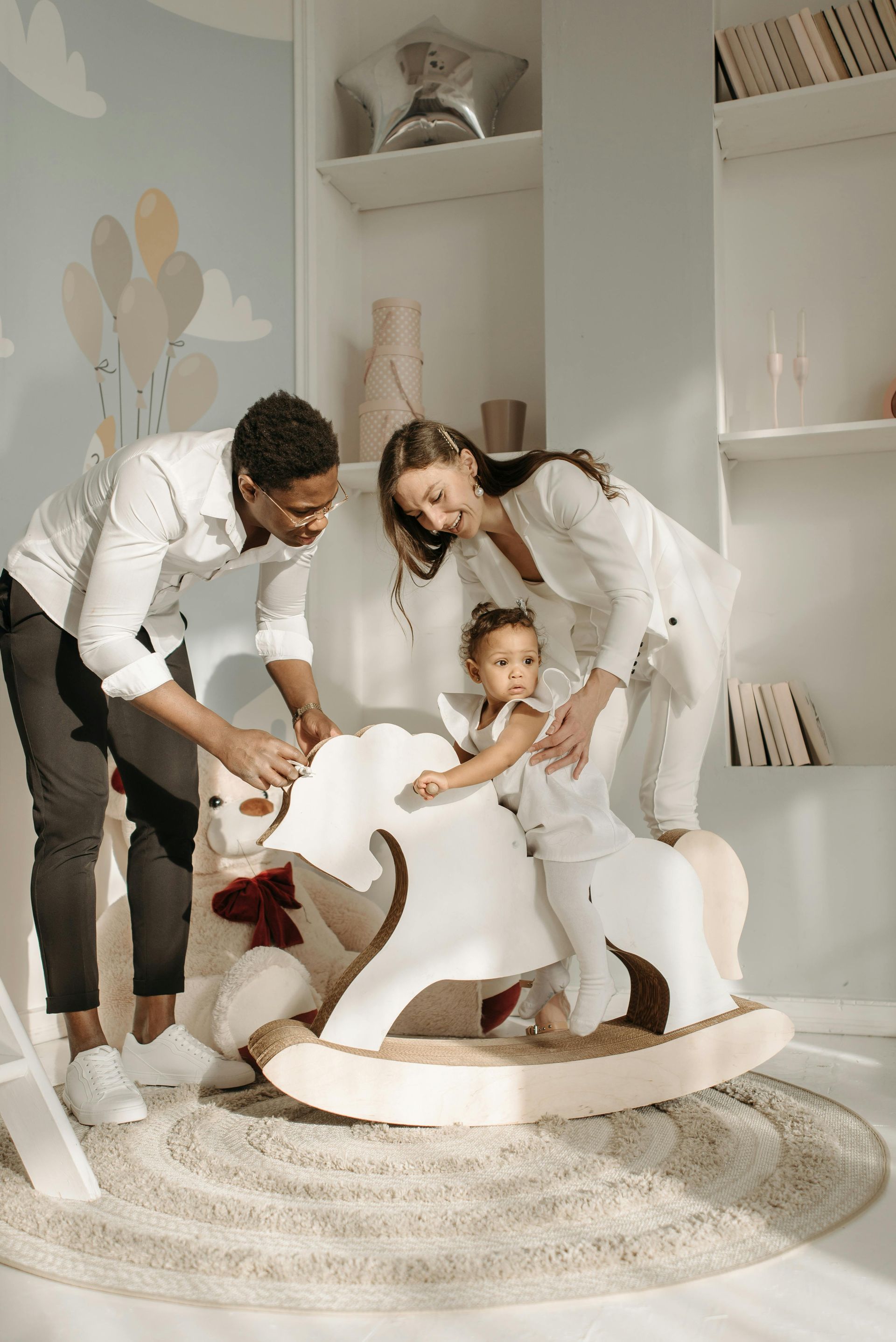 Family helping a child on a white rocking horse in a nursery, smiling.