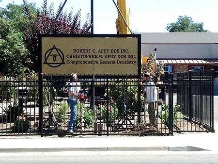 Church entrance with black iron gate and sign, people standing outside in bright daylight.