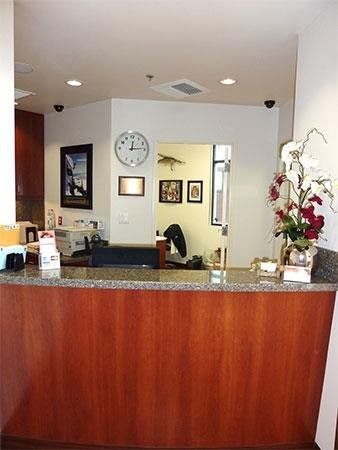Reception desk with granite counter, wood paneling, clock, wall art, and flowers in a clinic lobby