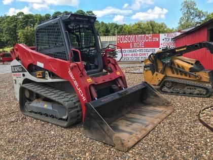 Red and yellow skid steer loaders parked on a gravel lot near a sign and red building.
