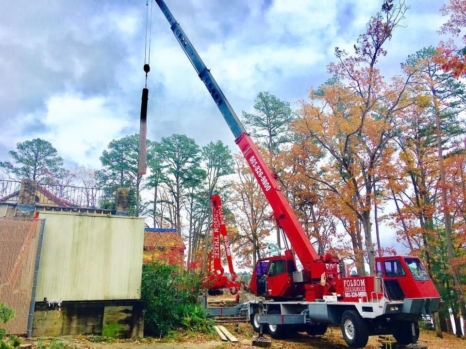 Crane lifting a pole beside a building, with autumn trees and a red truck in the background.