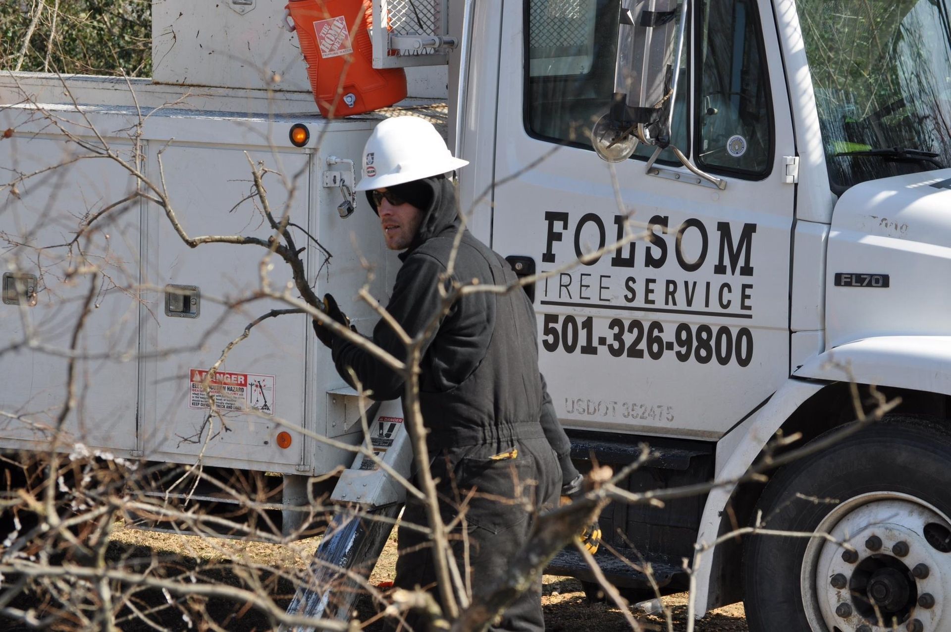 Worker in white hard hat beside a white tree service truck in a wooded area.