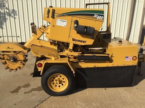 Yellow Vermeer stump grinder parked beside a building, with cutting wheel exposed.