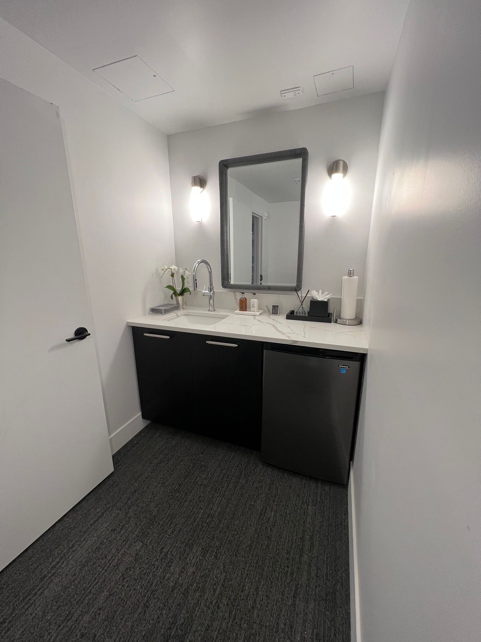 Small bathroom with black cabinets, white countertop, mirror, and a small refrigerator.