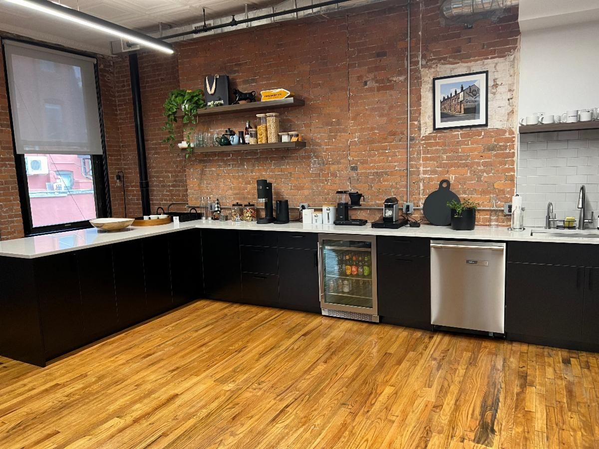 A kitchen with black cabinets, brick wall, wooden floor, and stainless steel appliances.