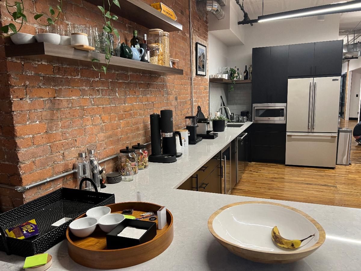 Kitchen with brick wall, shelves, appliances, and counter with fruit and snacks.