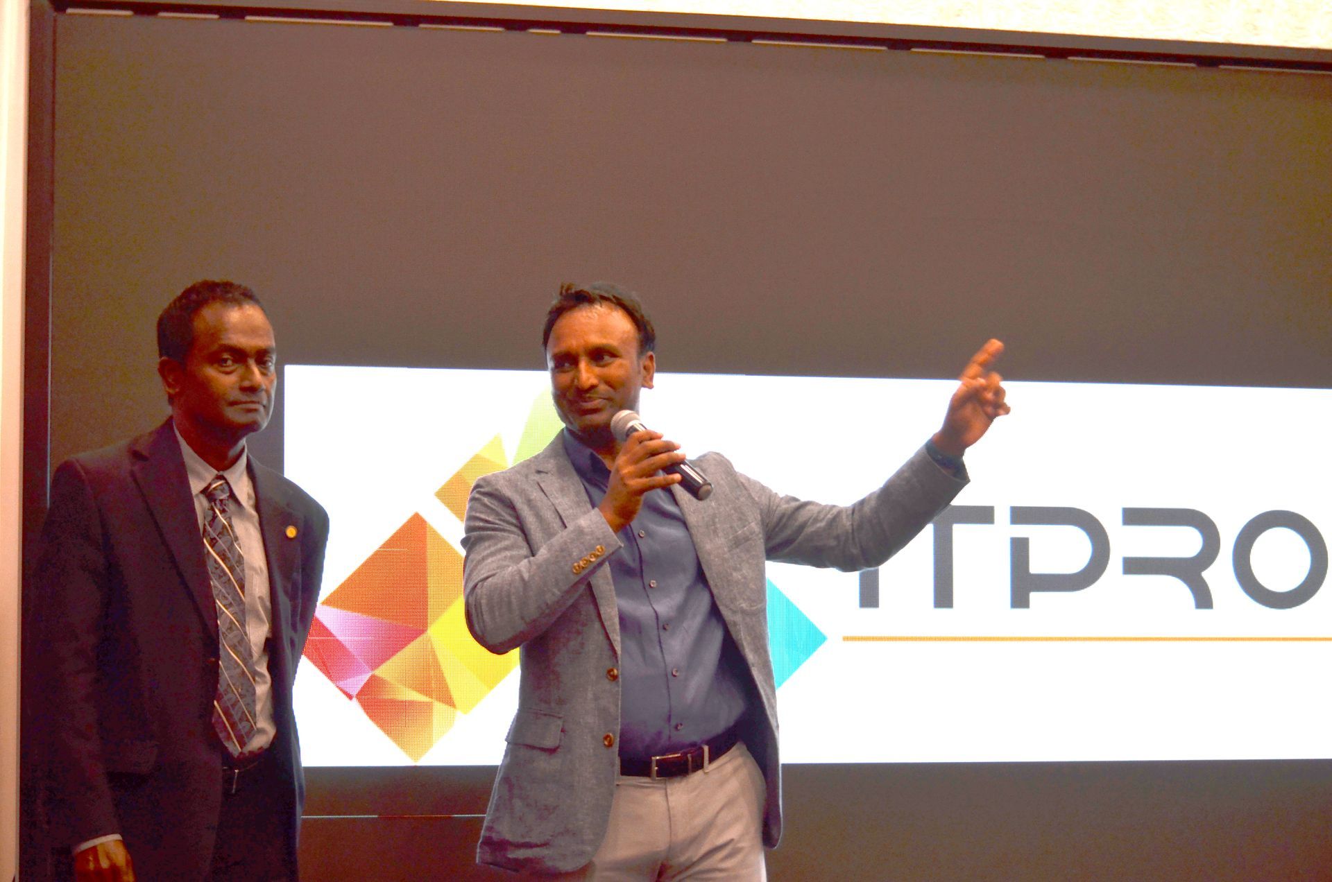 Two men speaking on stage in front of a lit presentation screen with the word “ITPRO” visible.