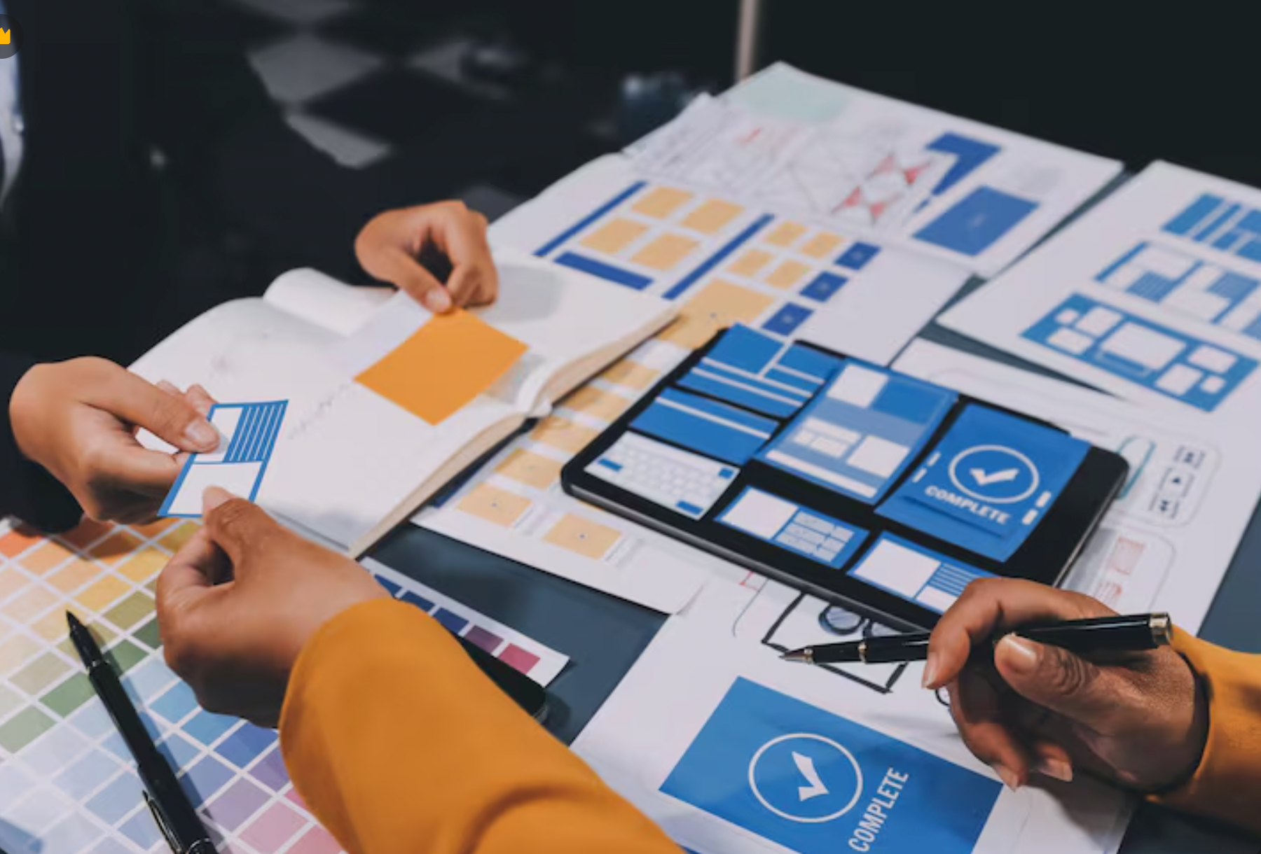 Hands reviewing blue and orange UI mockups on a desk with laptop and sketches