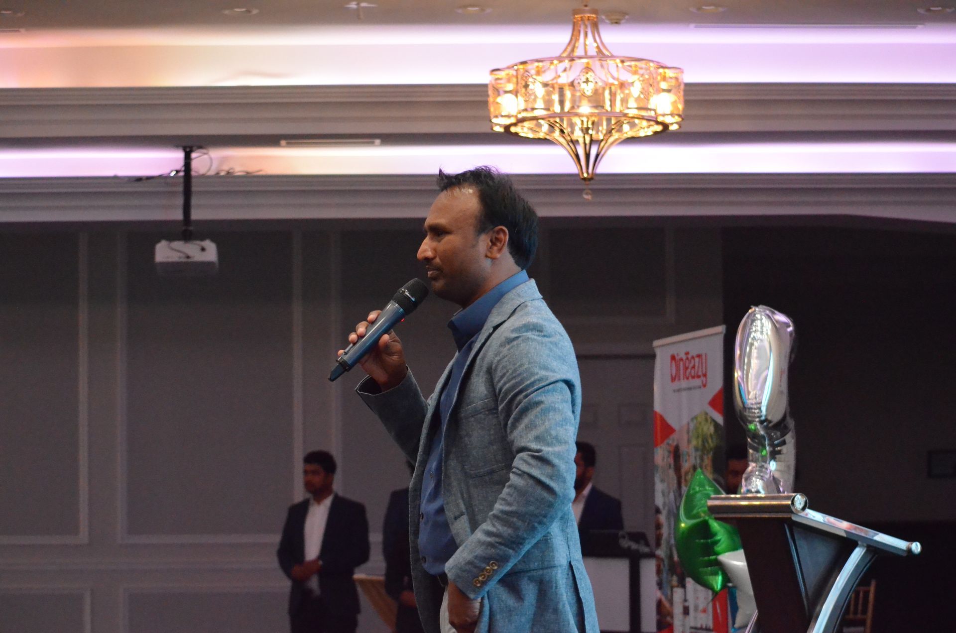 Man speaking into a microphone at an indoor event, with a man standing in the background and a chandelier overhead