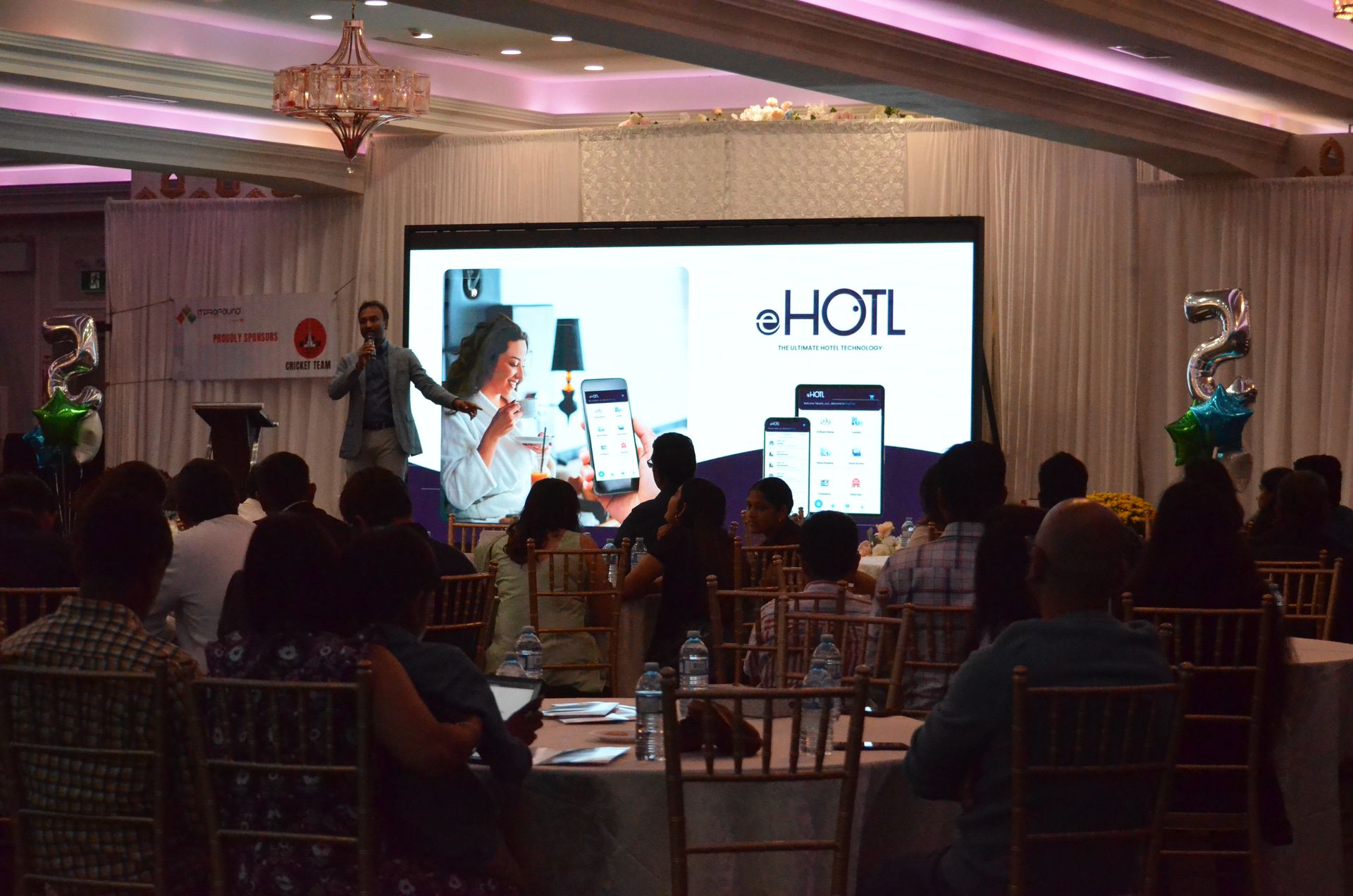 Conference presentation in a banquet hall with a large screen and seated audience in front of a stage.