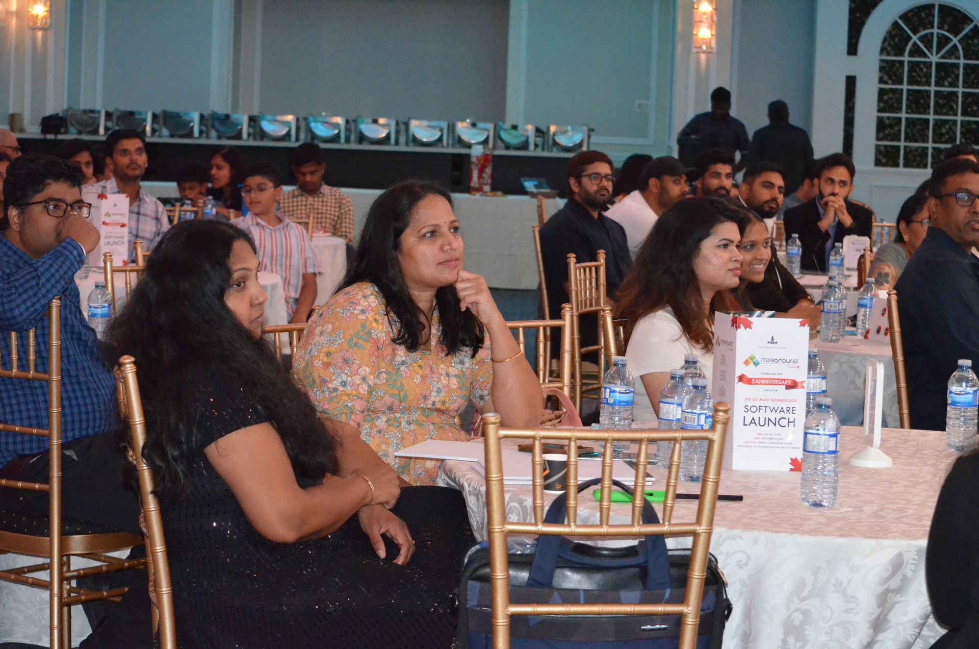 People seated at banquet tables in a formal event hall, listening attentively.