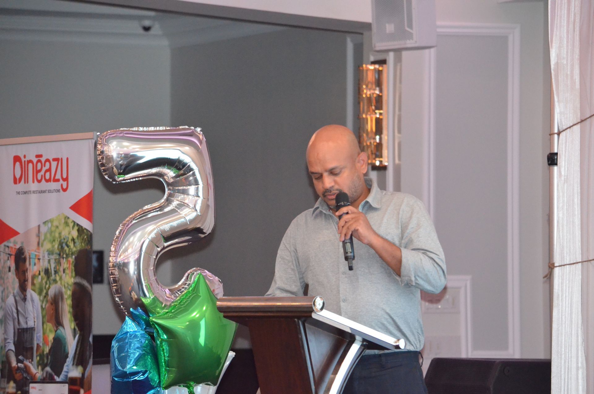 Man speaking at a podium with a microphone, beside silver and green balloon decorations.