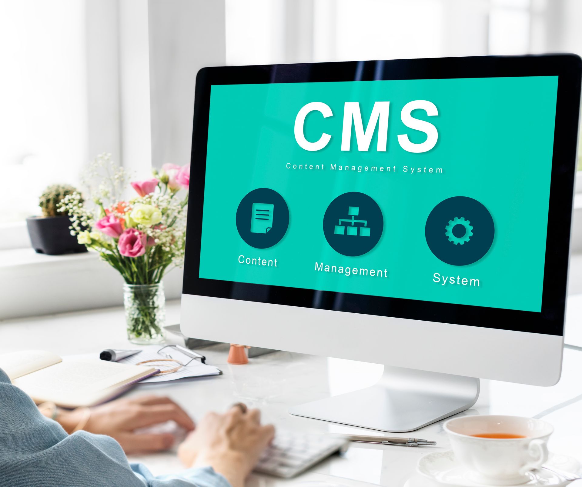 Person using desktop computer displaying CMS dashboard on teal screen in a bright office.