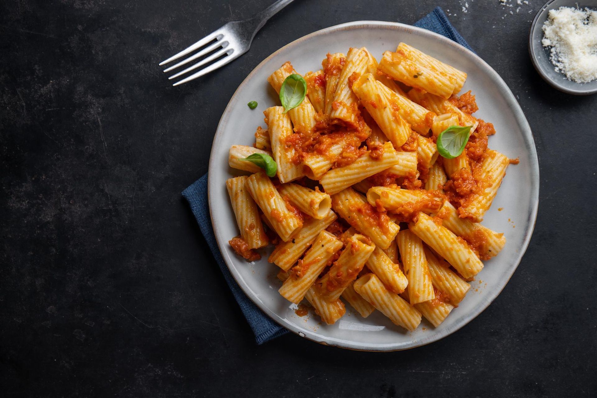 Plato de pasta rigatoni con salsa roja, guarnición de albahaca, queso y un tenedor sobre una superficie oscura.