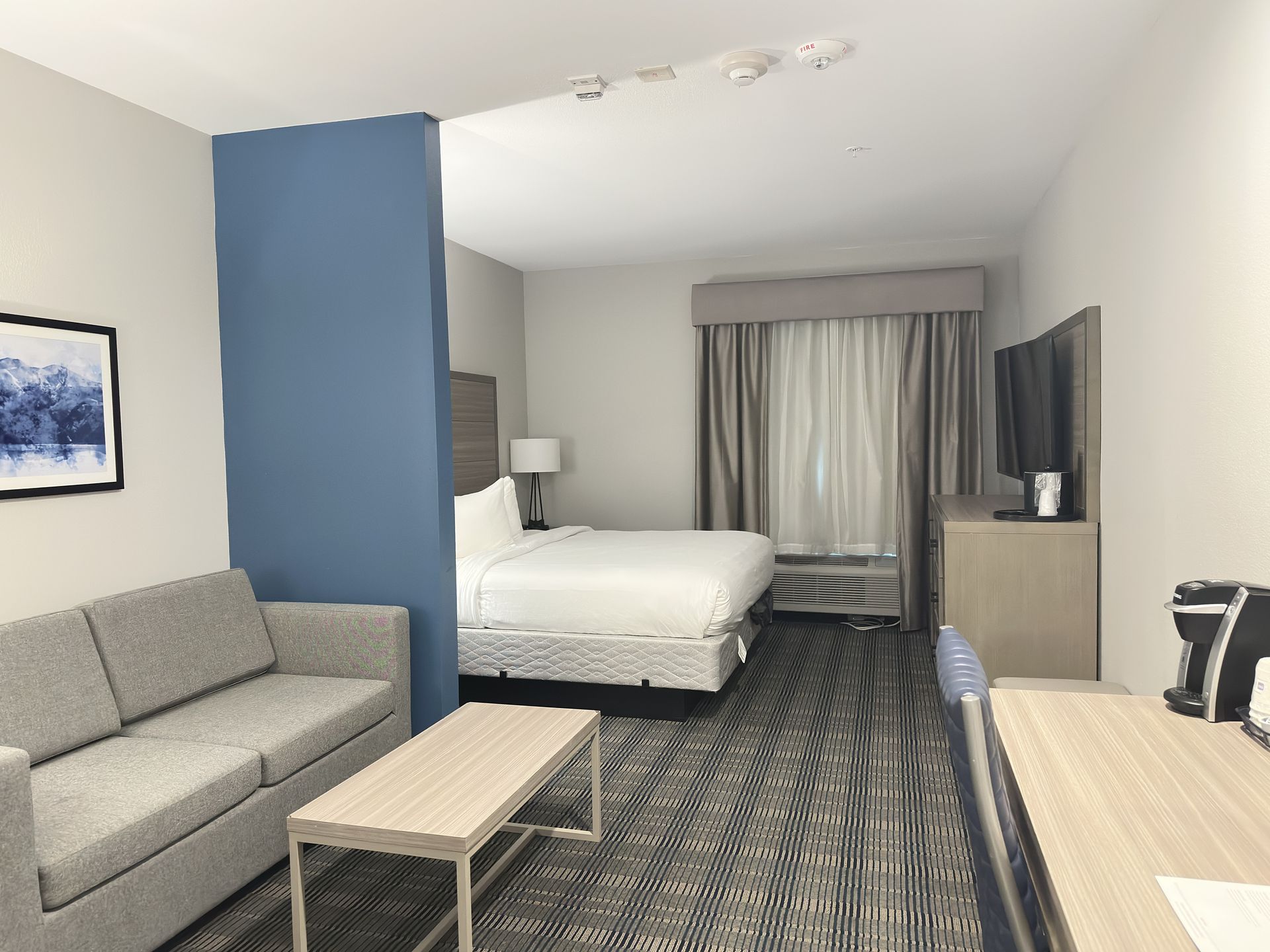 Hotel room with a bed, sofa, desk, and TV; a blue wall separates the living area.