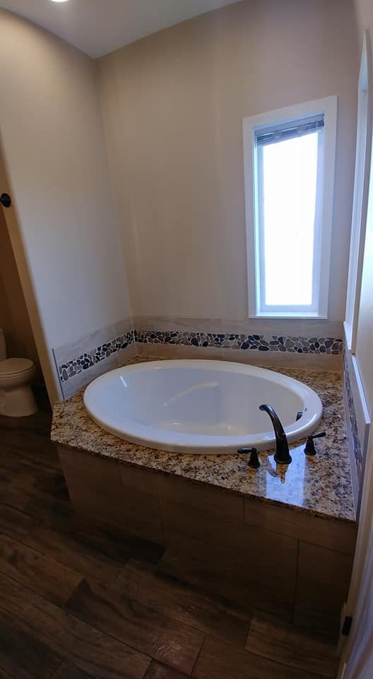 Bathroom corner with a built-in jetted tub, granite surround, wood flooring, and a narrow window.