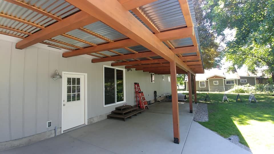 Covered patio beside a white house, with wooden posts, concrete floor, and a grassy yard.