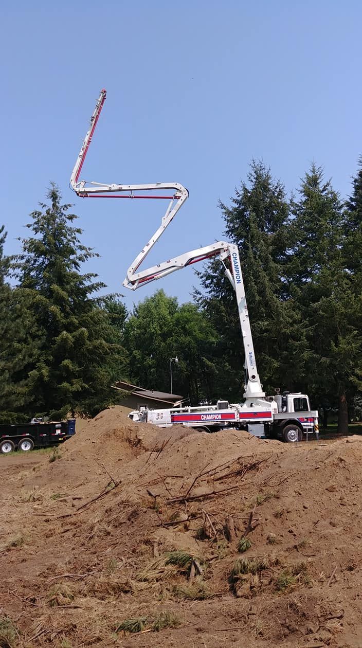 Concrete pump truck extending a zigzag boom over a dirt hillside beside trees and a road