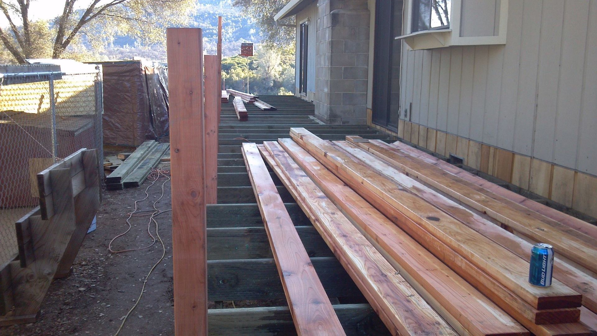 Stacked lumber beside a house, with a blue can on top, and a partially built wooden deck frame.