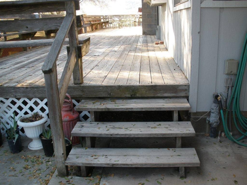 Wooden steps leading to a weathered deck beside a house, with potted plants and a green hose nearby