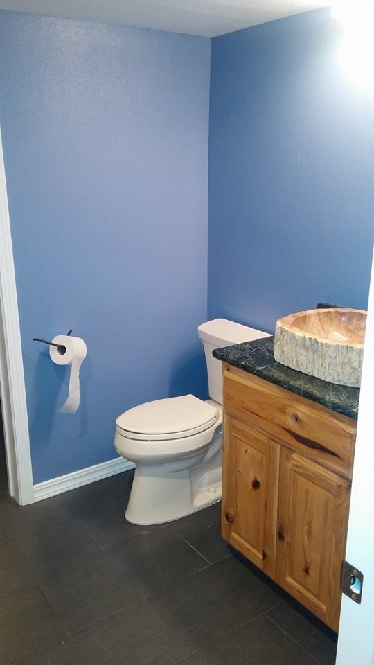 Small bathroom with blue walls, toilet, toilet paper holder, and wood vanity with black countertop.