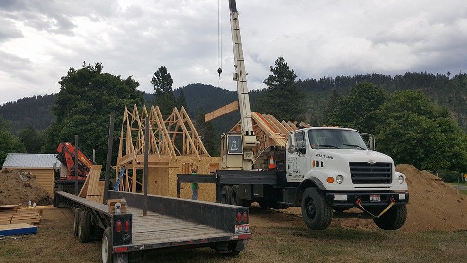 Construction site with a truck-mounted crane lifting roof trusses beside a flatbed trailer.