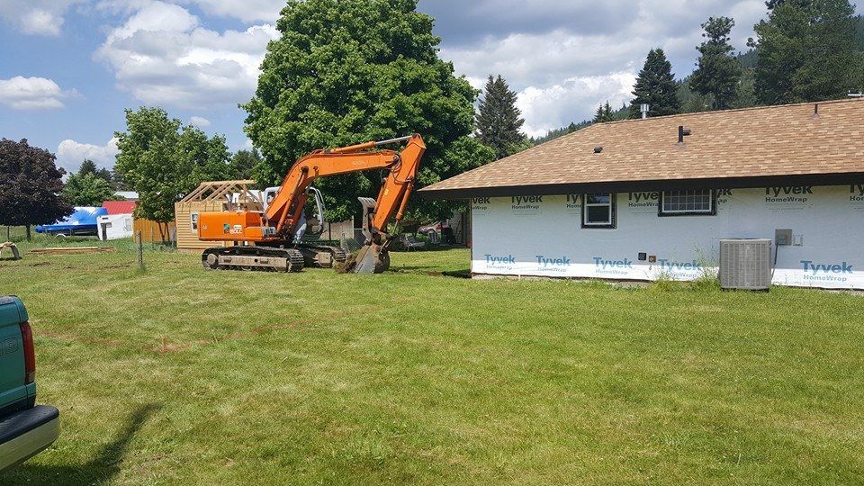Orange excavator beside a light blue house on a grassy lawn under a partly cloudy sky