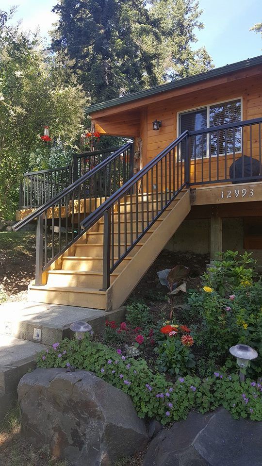 Wooden house exterior with black-railed stairs, flower beds, and trees in a landscaped yard