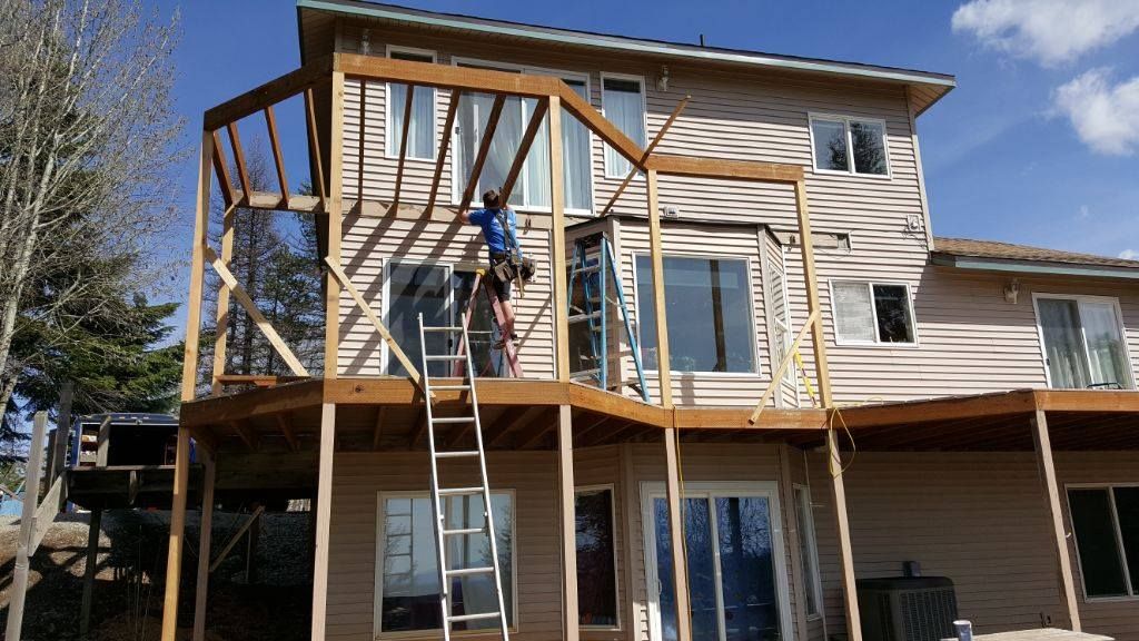 Person working on a wooden deck railing on a two-story house, with a ladder and scaffolding.