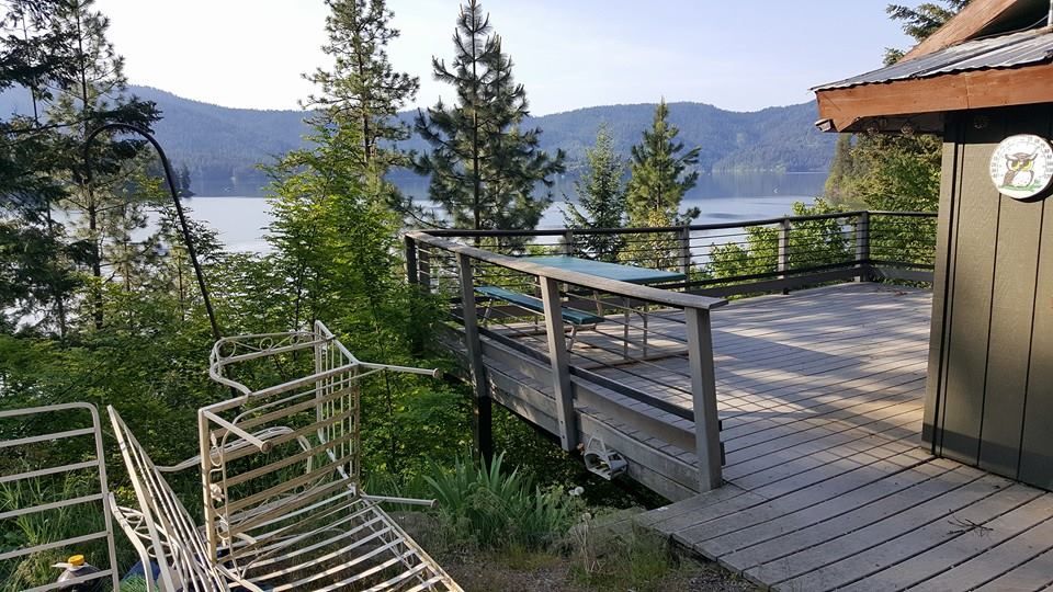 Wooden deck with railing and chairs overlooking a lake and mountains, beside a small cabin