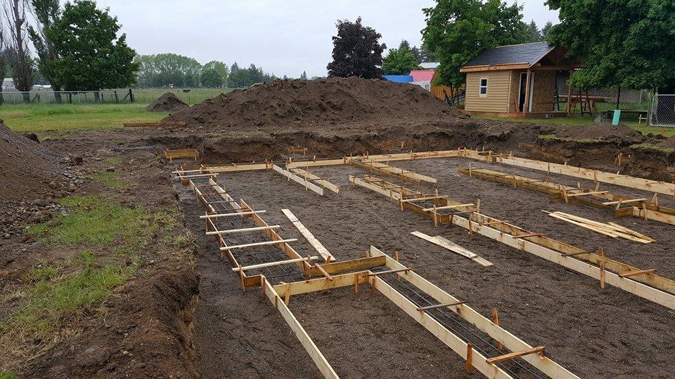 Foundation framing on a dirt lot with wooden forms and soil piles near a small house in overcast weather