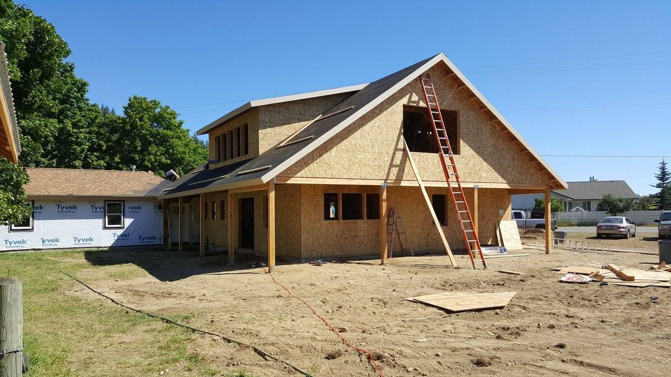 House under construction with workers on scaffolding and a ladder in front of a partially finished wooden roof.