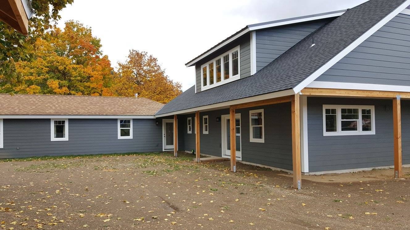 Gray house with dark roof and autumn trees, seen from a courtyard with a covered porch.