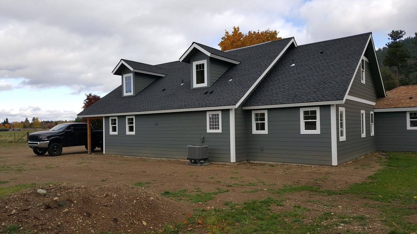 Gray farmhouse with dark roof and dormers in a rural yard, with a black truck parked nearby.