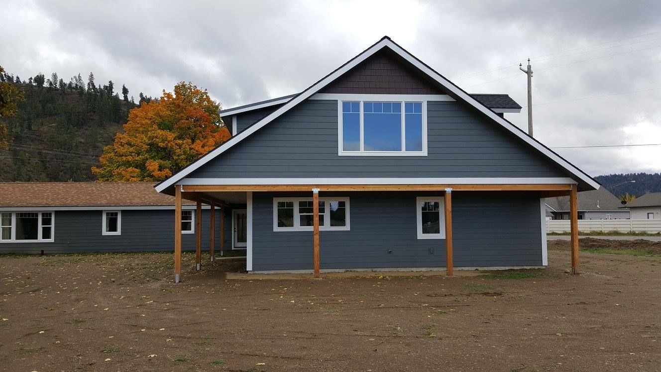 Blue-gray house with a dark roof and large front windows on a gravel lot, with autumn trees behind.