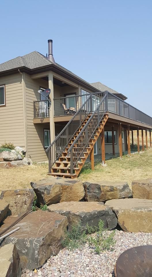 Two-story beige house with exterior wooden stairs and a deck, set on a rocky hillside under a blue sky