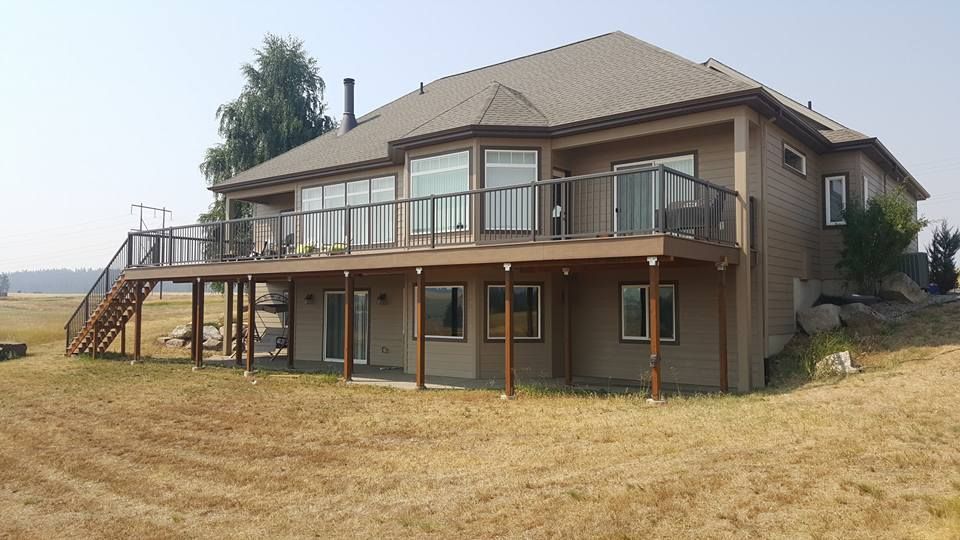 Two-story tan house with a wraparound deck and stairs on a dry grassy lot