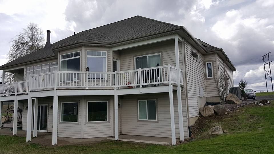Two-story beige house with white trim, wraparound porch, and stone foundation on a grassy lot.